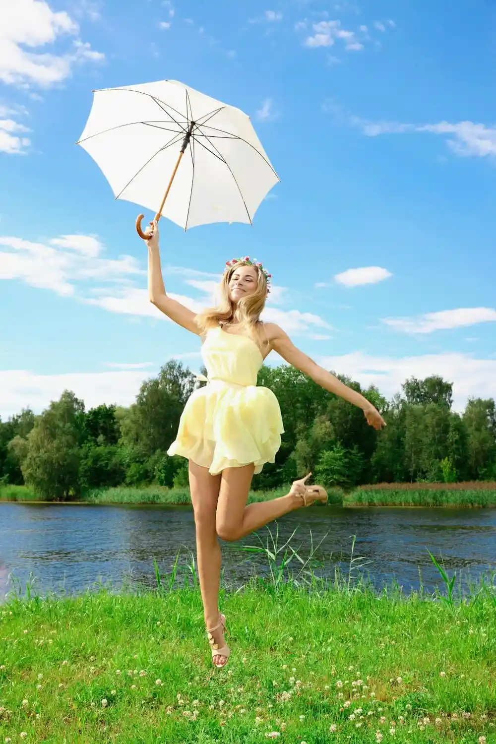 Vrolijke vrouw in gele zomerjurk met witte parasol, springend bij een schilderachtig meer